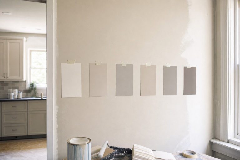 Several plain paint color sample cards taped in a row on a wall for comparison
