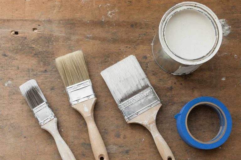 Overhead view of three paint brushes, an open can of off-white paint, and a roll of painters tape on a wooden workbench