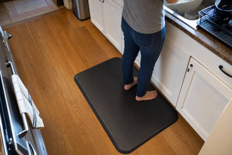Looking down at a person's feet on a cushioned anti-fatigue mat laid over hardwood in front of a kitchen sink