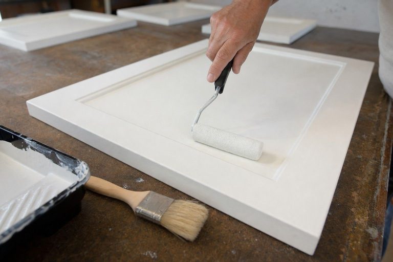A hand rolling a small foam roller across a cabinet door, leaving a flat even coat with no roller texture